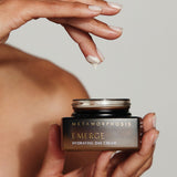A person holding an open Metamorphosis Emerge cream jar in one hand while lifting a small dab of cream from it on a fingertip.