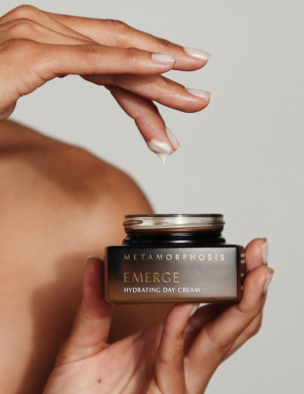 A person holding an open Metamorphosis Emerge cream jar in one hand while lifting a small dab of cream from it on a fingertip.