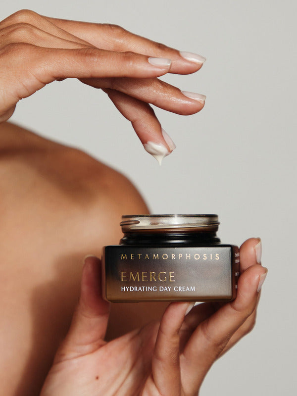 A person holding an open Metamorphosis Emerge cream jar in one hand while lifting a small dab of cream from it on a fingertip.