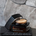 Open dark amber Metamorphosis Evolve cream jar on a rough black slate tile, showing pale cream inside, with its black lid propped beside it against a grey stone wall.