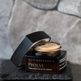 Open dark amber Metamorphosis Evolve cream jar on a rough black slate tile, showing pale cream inside, with its black lid propped beside it against a grey stone wall.