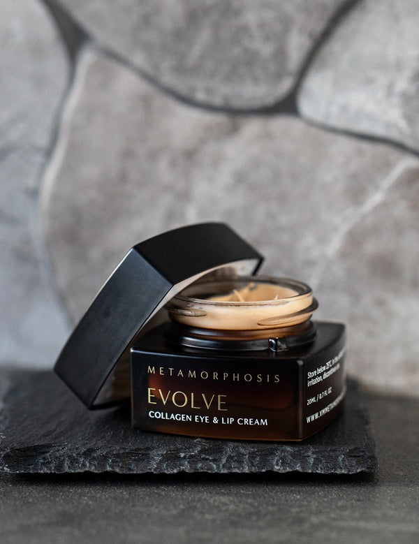 Open dark amber Metamorphosis Evolve cream jar on a rough black slate tile, showing pale cream inside, with its black lid propped beside it against a grey stone wall.