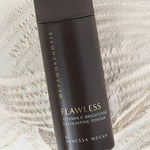 Tall dark brown Metamorphosis Flawless exfoliating-powder container with gold lettering lying diagonally over fine pale powder raked into curved ridges.