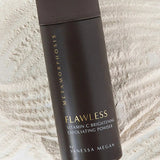 Tall dark brown Metamorphosis Flawless exfoliating-powder container with gold lettering lying diagonally over fine pale powder raked into curved ridges.