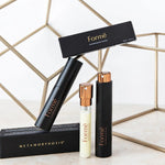 Slim black Formë perfume sprays and a pale refill vial with gold spray tops arranged with black cartons in front of gold wire-frame cubes.