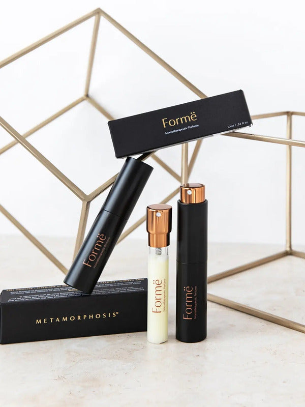 Slim black Formë perfume sprays and a pale refill vial with gold spray tops arranged with black cartons in front of gold wire-frame cubes.