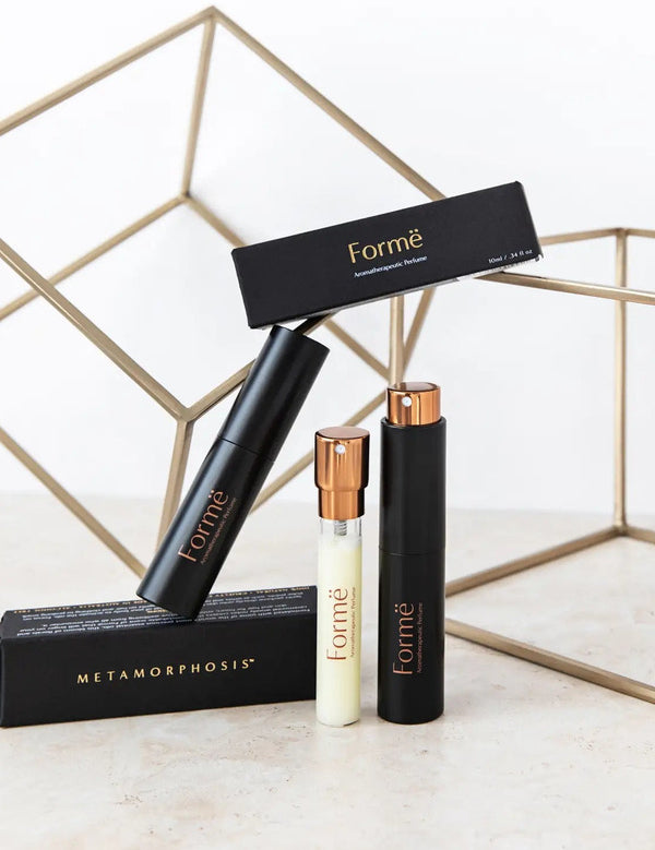 Slim black Formë perfume sprays and a pale refill vial with gold spray tops arranged with black cartons in front of gold wire-frame cubes.