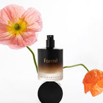 Black Metamorphosis Formë Aromatherapeutic Perfume bottle and round cap displayed between soft pink and orange poppy flowers on white.