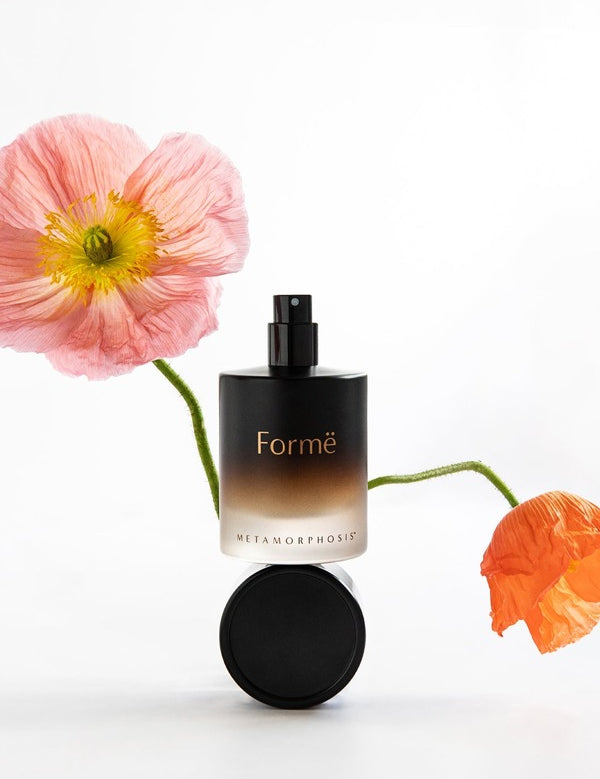 Black Metamorphosis Formë Aromatherapeutic Perfume bottle and round cap displayed between soft pink and orange poppy flowers on white.