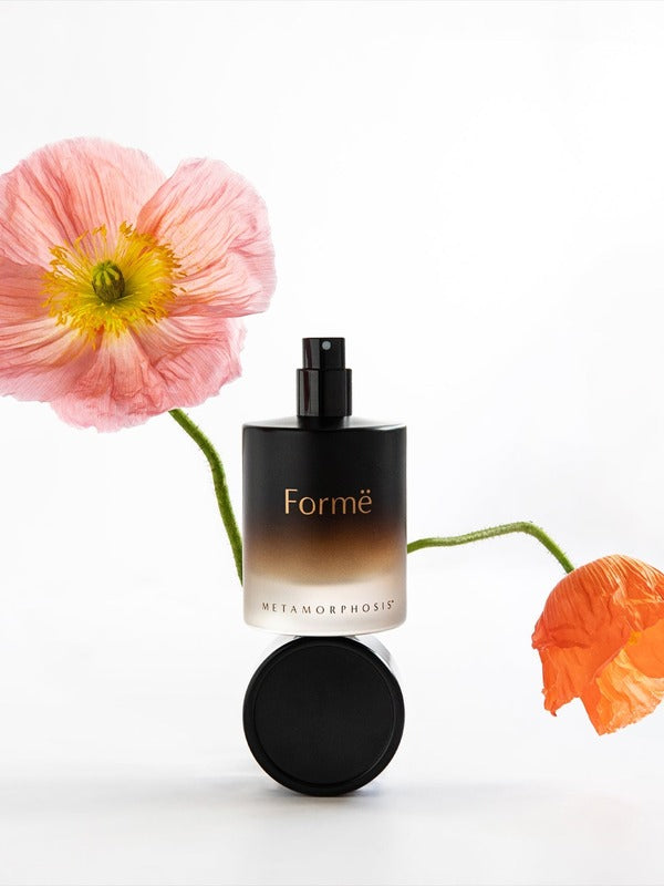 Black Metamorphosis Formë Aromatherapeutic Perfume bottle and round cap displayed between soft pink and orange poppy flowers on white.