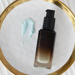 Dark amber Metamorphosis Illuminate pump bottle beside a pale blue cream smear on a round gold-rimmed mirror resting on a white ridged surface.