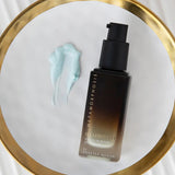 Dark amber Metamorphosis Illuminate pump bottle beside a pale blue cream smear on a round gold-rimmed mirror resting on a white ridged surface.