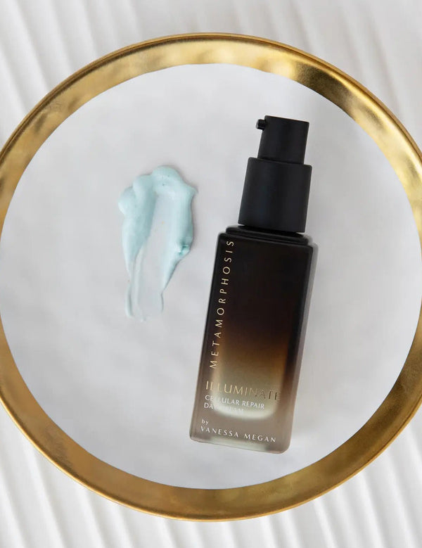 Dark amber Metamorphosis Illuminate pump bottle beside a pale blue cream smear on a round gold-rimmed mirror resting on a white ridged surface.