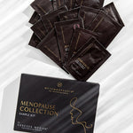 Black Metamorphosis Menopause Collection Starter Kit box below a fan of dark sample sachets on white marble with diagonal light and shadow.