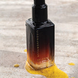 Metamorphosis Re-Gen Cell Repair Oil bottle standing on pale stone beside a textured sweep of golden powder.