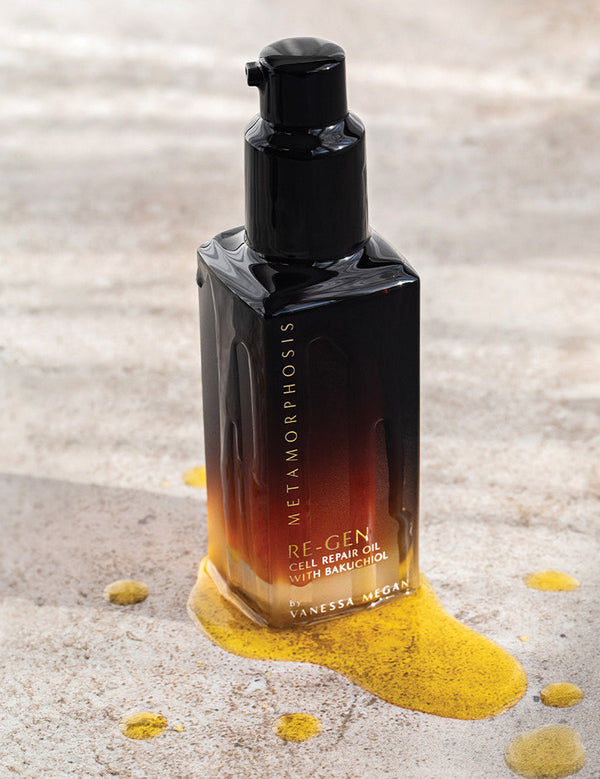 Metamorphosis Re-Gen Cell Repair Oil bottle standing on pale stone beside a textured sweep of golden powder.