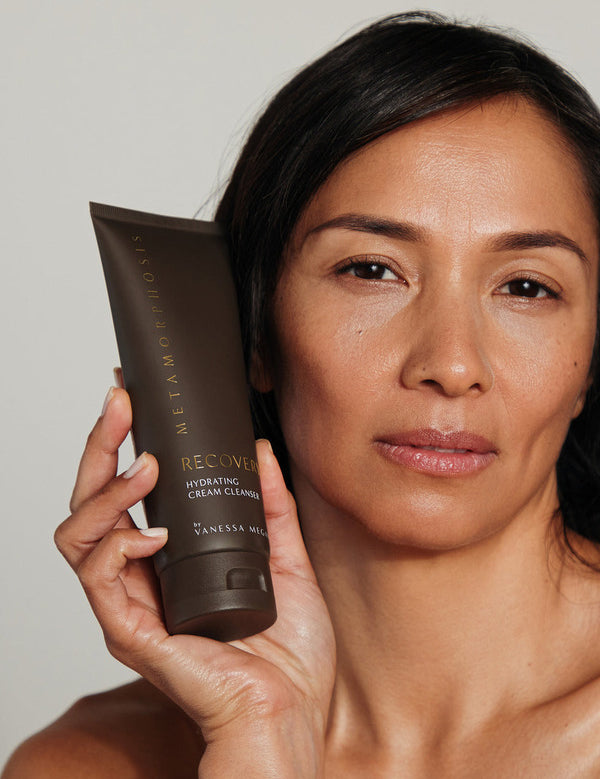 Woman holding a tube of Metamorphosis Recovery Hydrating Cream Cleanser beside her face against a softly lit neutral background.