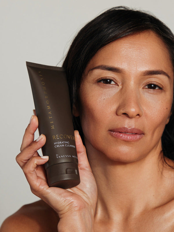 Woman holding a tube of Metamorphosis Recovery Hydrating Cream Cleanser beside her face against a softly lit neutral background.