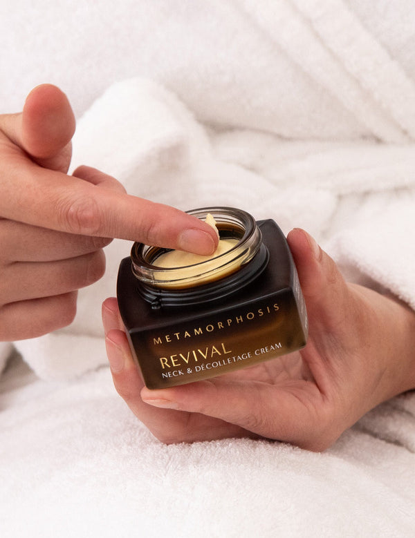 Person in a white bathrobe holding an open Metamorphosis Revival cream jar while touching the pale cream inside with a fingertip.