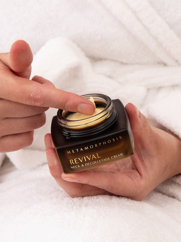 Person in a white bathrobe holding an open Metamorphosis Revival cream jar while touching the pale cream inside with a fingertip.