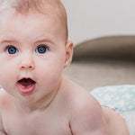 Baby lying on their tummy with their head raised and mouth open, looking towards the camera on a pale patterned blanket.