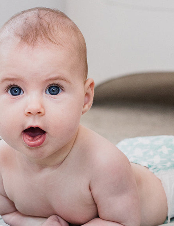 Baby lying on their tummy with their head raised and mouth open, looking towards the camera on a pale patterned blanket.