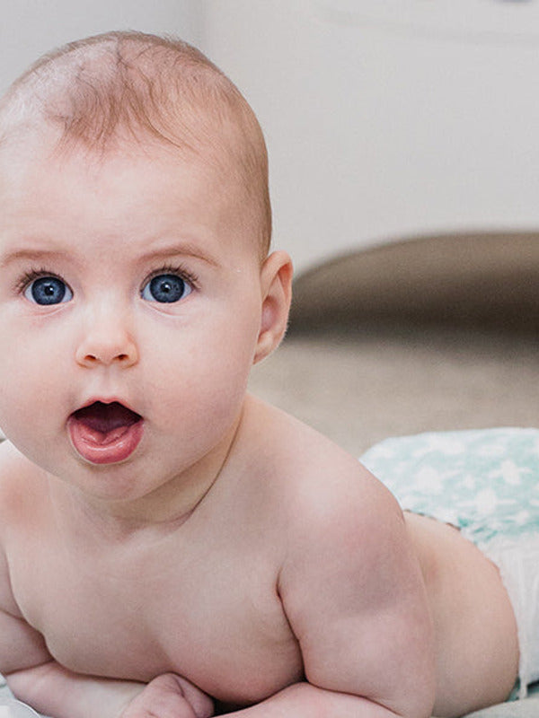 Baby lying on their tummy with their head raised and mouth open, looking towards the camera on a pale patterned blanket.