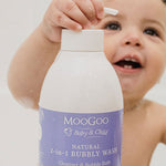 Smiling young child in a bath holding a large white MooGoo Bubbly Wash pump bottle with a purple label, with one hand on the pump.