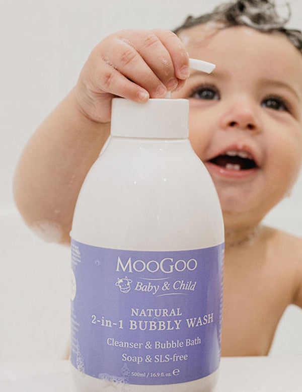 Smiling young child in a bath holding a large white MooGoo Bubbly Wash pump bottle with a purple label, with one hand on the pump.