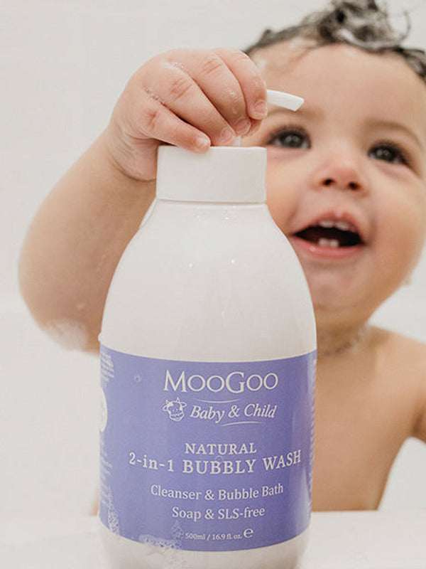 Smiling young child in a bath holding a large white MooGoo Bubbly Wash pump bottle with a purple label, with one hand on the pump.