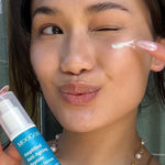 Person winking while dabbing white serum beneath one eye with a fingertip and holding a blue-labelled MooGoo Amplified Peptide Active Serum bottle.