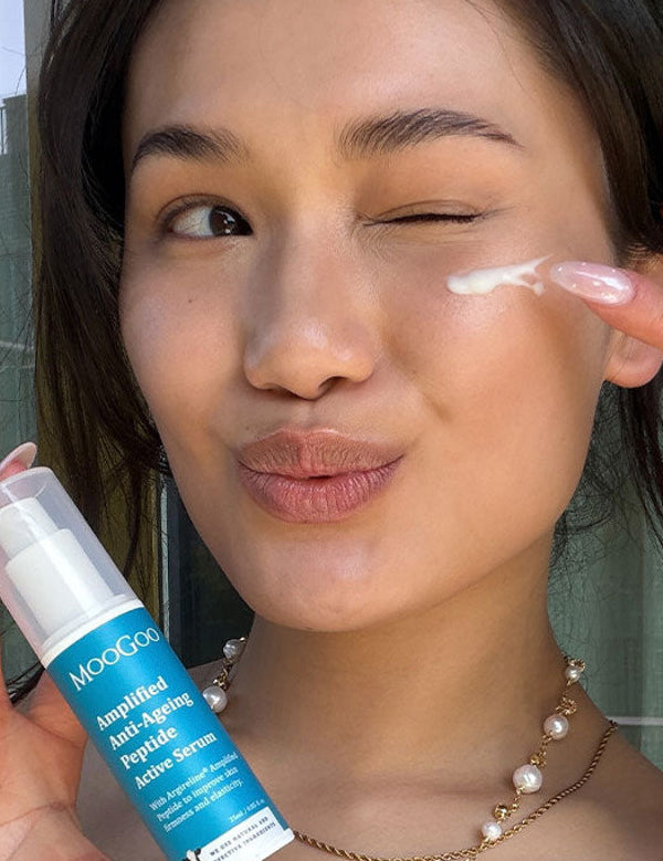 Person winking while dabbing white serum beneath one eye with a fingertip and holding a blue-labelled MooGoo Amplified Peptide Active Serum bottle.