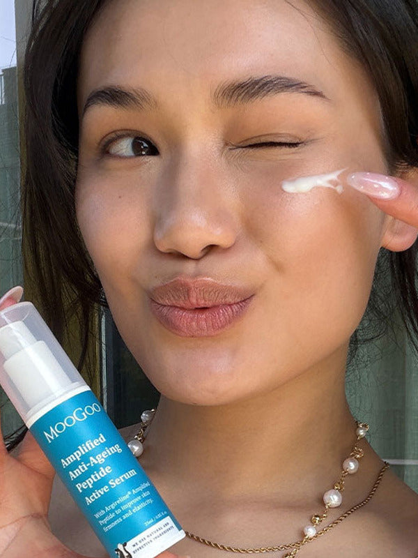 Person winking while dabbing white serum beneath one eye with a fingertip and holding a blue-labelled MooGoo Amplified Peptide Active Serum bottle.