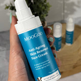 Hand holding a white MooGoo Anti-Ageing Skin Renewal Face Cream pump bottle with a blue label, with two other bottles on a wooden tray behind.