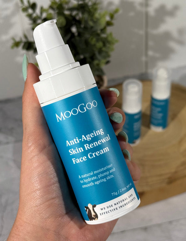 Hand holding a white MooGoo Anti-Ageing Skin Renewal Face Cream pump bottle with a blue label, with two other bottles on a wooden tray behind.
