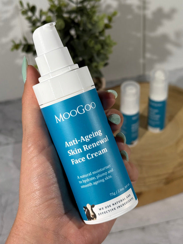 Hand holding a white MooGoo Anti-Ageing Skin Renewal Face Cream pump bottle with a blue label, with two other bottles on a wooden tray behind.