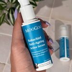 Hand holding an uncapped MooGoo Bakuchiol Anti-Ageing Active Serum pump bottle with a blue label, with another bottle and green leaves behind.