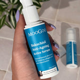 Hand holding an uncapped MooGoo Bakuchiol Anti-Ageing Active Serum pump bottle with a blue label, with another bottle and green leaves behind.