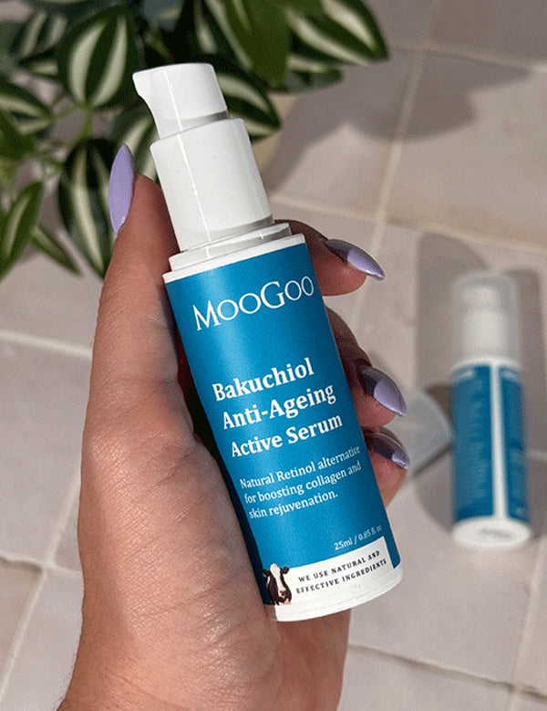 Hand holding an uncapped MooGoo Bakuchiol Anti-Ageing Active Serum pump bottle with a blue label, with another bottle and green leaves behind.