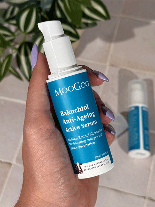 Hand holding an uncapped MooGoo Bakuchiol Anti-Ageing Active Serum pump bottle with a blue label, with another bottle and green leaves behind.