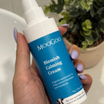 Hand with lilac nail polish holding a blue-labelled MooGoo Blemish Calming Cream 75g pump bottle beside a white basin and leafy plant.