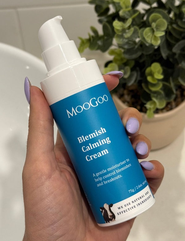 Hand with lilac nail polish holding a blue-labelled MooGoo Blemish Calming Cream 75g pump bottle beside a white basin and leafy plant.