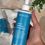 Hand holding a MooGoo Creamy Hydrating Face Cleanser pump bottle above a white towel with another bottle and trailing greenery.