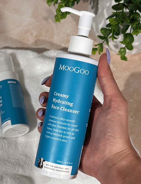 Hand holding a MooGoo Creamy Hydrating Face Cleanser pump bottle above a white towel with another bottle and trailing greenery.