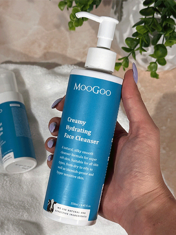 Hand holding a MooGoo Creamy Hydrating Face Cleanser pump bottle above a white towel with another bottle and trailing greenery.