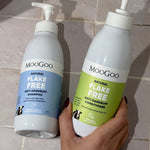 Hand holding a green-labelled MooGoo Flake Free conditioner pump bottle beside the blue-labelled shampoo bottle on beige bathroom tiles.