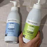 Hand holding a green-labelled MooGoo Flake Free conditioner pump bottle beside the blue-labelled shampoo bottle on beige bathroom tiles.
