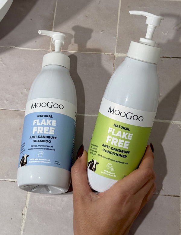 Hand holding a green-labelled MooGoo Flake Free conditioner pump bottle beside the blue-labelled shampoo bottle on beige bathroom tiles.