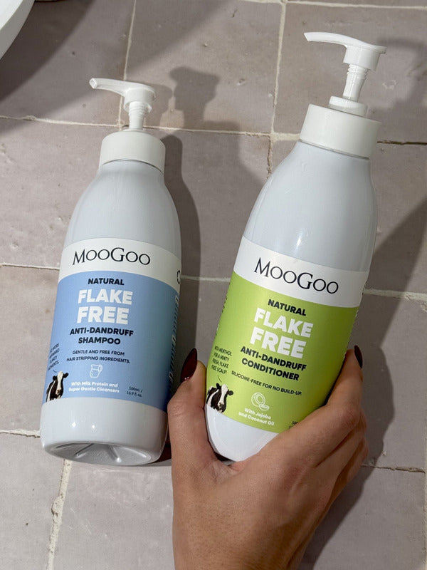 Hand holding a green-labelled MooGoo Flake Free conditioner pump bottle beside the blue-labelled shampoo bottle on beige bathroom tiles.
