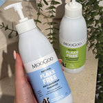 Hand holding a blue-labelled MooGoo Flake Free shampoo pump bottle, with the green-labelled conditioner and trailing foliage behind.